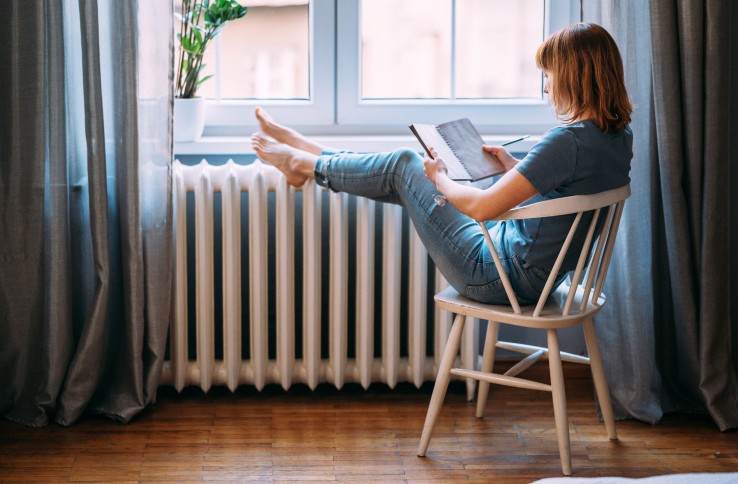 Une personne est assise près d'un radiateur, près de la fenêtre – image symbolique d'un chauffage respectueux du climat grâce à un système de chauffage au bois.