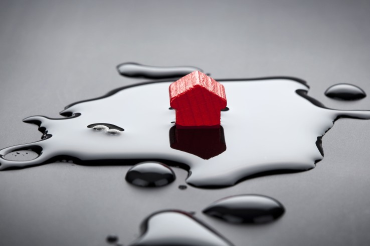 Une maquette de maison rouge se trouve dans une flaque d'eau, illustrant la protection d'une pompe à chaleur air-eau contre les inondations et le ruissellement.