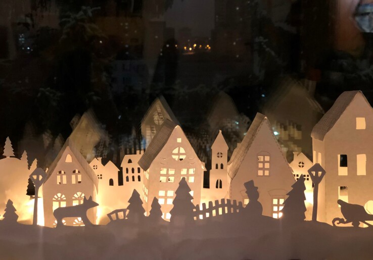 Fenêtre de l’Avent illuminée avec un décor hivernal de maisons et des figurines de Noël en papier.