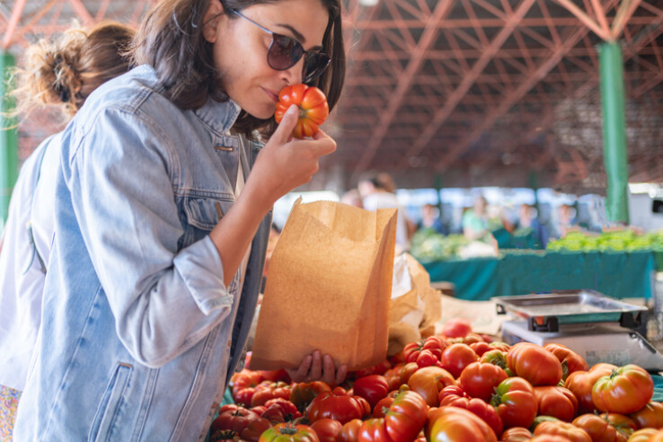 Une personne sent une tomate sur un marché hebdomadaire et achète des légumes frais dans un sac en papier.