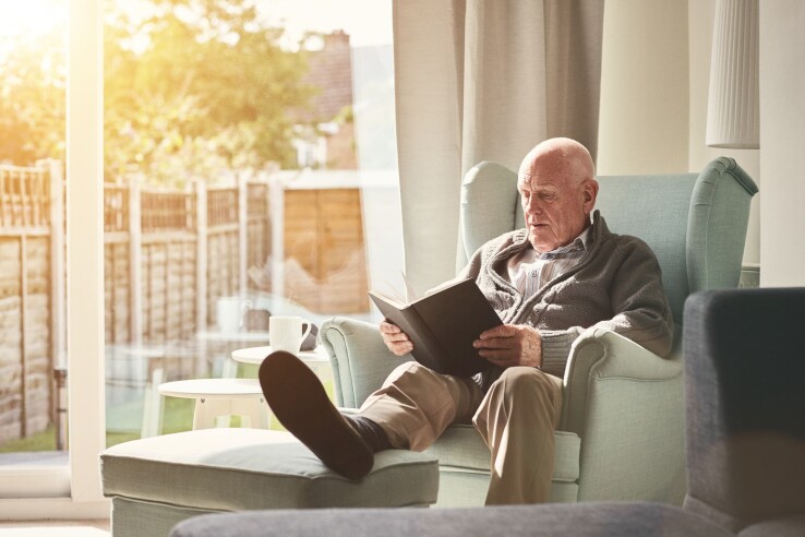 Un homme âgé est assis dans un salon lumineux et lit dans un fauteuil – un logement adapté aux personnes âgées.