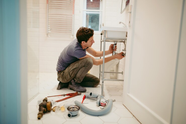 Une personne répare le siphon sous un lavabo de la salle de bains. Des outils et des pièces de rechange sont posés par terre : il s'agit d'une réparation domestique typique dans le domaine de la plomberie.