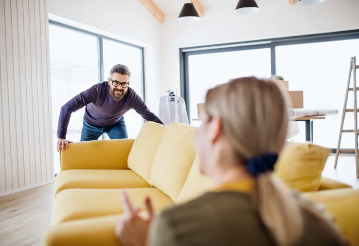 Un homme déplace un canapé jaune dans le salon, tandis qu'une femme l'observe.