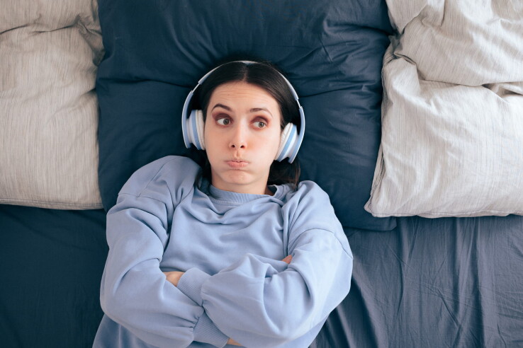 Une femme portant des écouteurs est allongée dans son lit, agacée, et illustre le problème des nuisances sonores dans une copropriété.