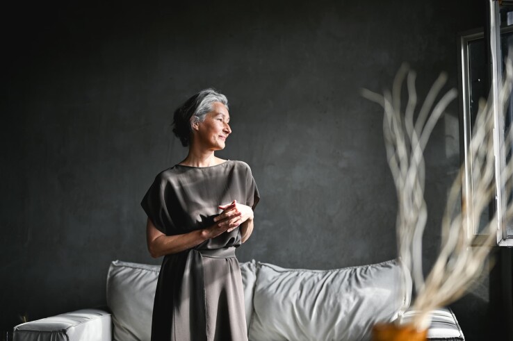 Ältere Frau in einem minimalistisch eingerichteten Wohnzimmer mit dunkler Wand und hellem Sofa.