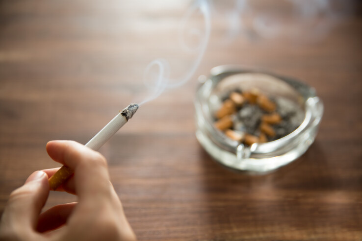 Une main tenant une cigarette allumée à côté d'un cendrier – image symbolique représentant le fait de fumer dans un appartement en location.