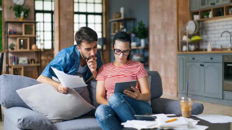 Un couple est assis sur un canapé, regarde une tablette et consulte des documents.