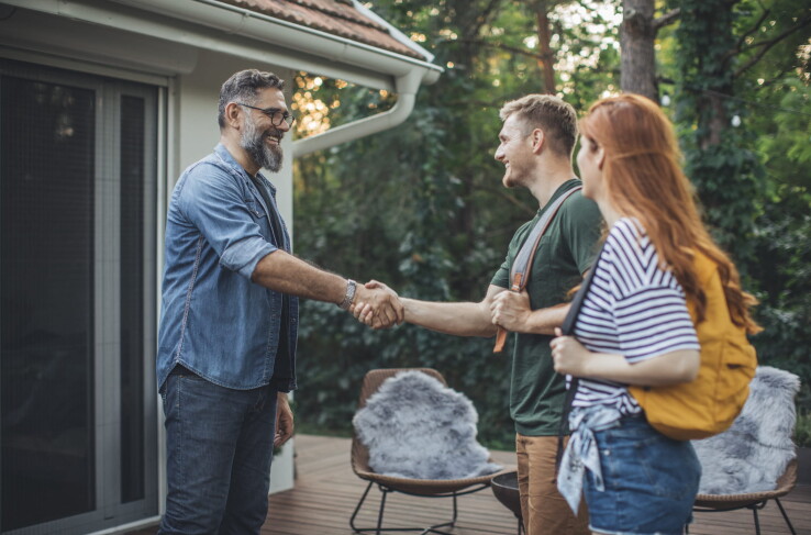 Un propriétaire accueille un couple devant une maison et discute avec eux de la location d'un appartement.