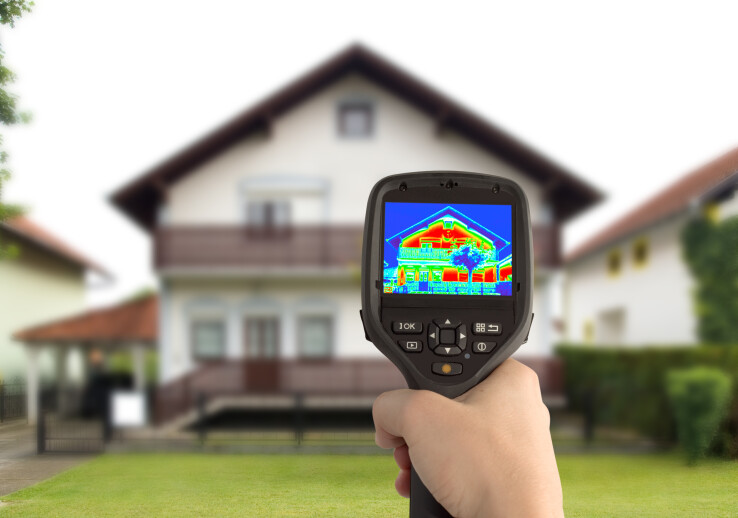 Une main tenant une caméra thermique devant une maison individuelle – image symbolique illustrant une analyse thermographique visant à détecter les déperditions thermiques et les ponts thermiques.