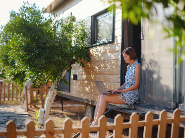 Une femme est assise devant une « tiny house » avec son ordinateur portable et travaille dans l'entrée de cette habitation compacte.