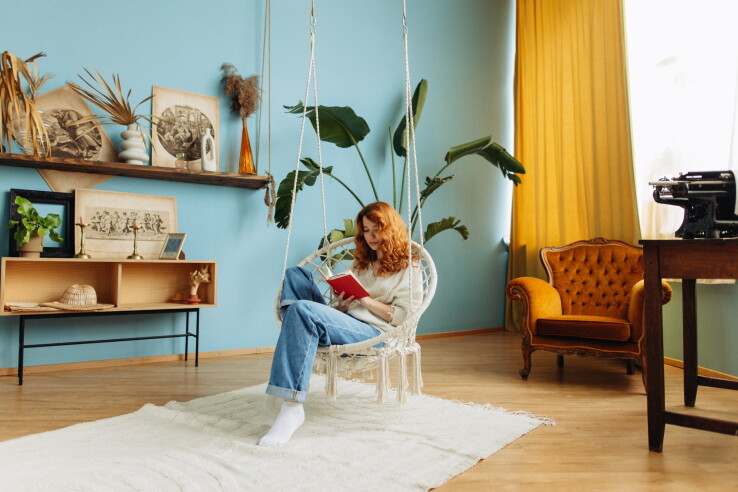 Une femme lit un livre dans une chaise suspendue, dans son appartement aux couleurs vives.